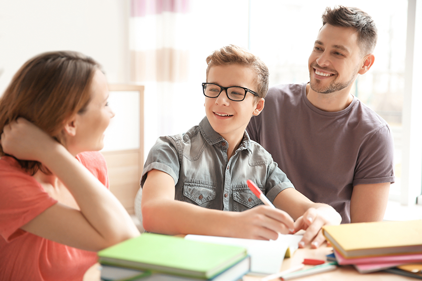 Jongen met pen boven schrift kijkt blij naar vrouw naast hem, man zit erachter | De VO Gids 2027
