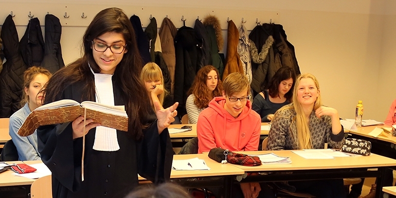 Themascholen: Pre university als themaschool. Meisje in toga leest voor uit leerboek met andere leerlingen in klaslokaal De VO Gids 2026