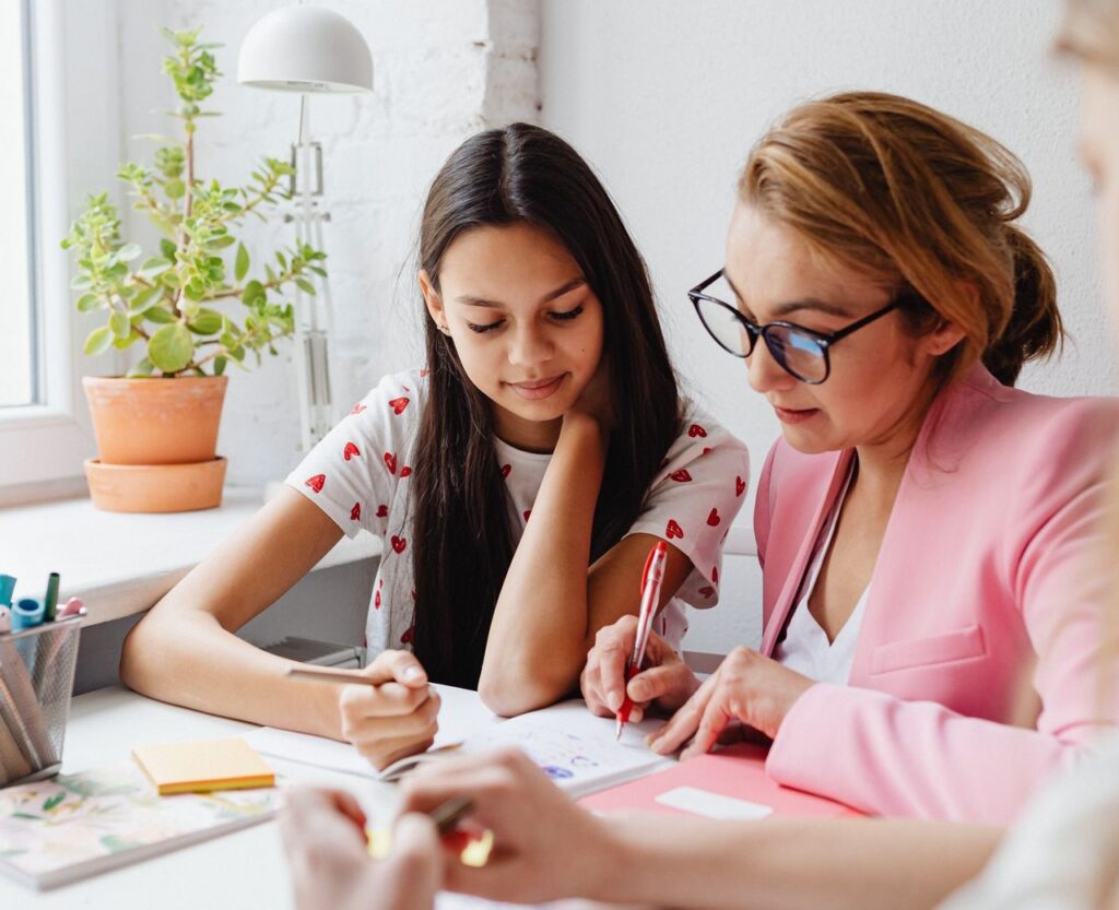 Vrouw in roze jasje zit naast meisje aan tafel voor een schrift | De VO Gids 2027
