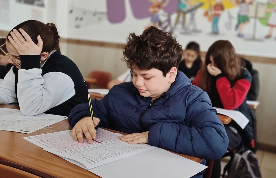 Jongen met jas aan vult toetspapier in met potlood, naast hem een meisje dsat haar hoofd ondersteunt | De VO Gids 2027