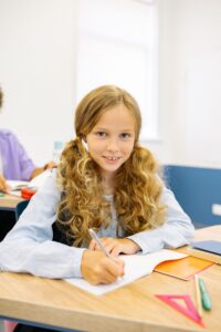 Meisje met staarten lacht naar de camera terwijl ze met pen in hand boven schrift zit