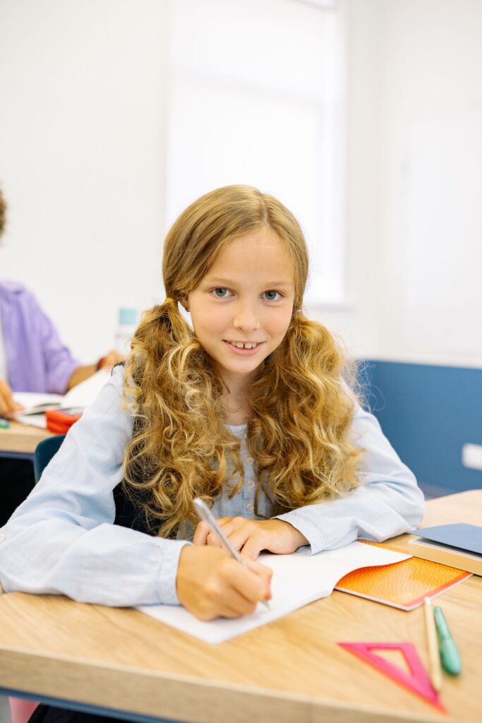 Meisje met staarten lacht naar de camera terwijl ze met pen in hand boven schrift zit